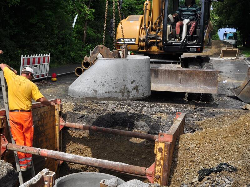 Ein Arbeiter auf einer Baustelle überwacht einen Bagger, der ein großes Betonrohr in ein Erdloch absenkt, umgeben von grünem Laub.