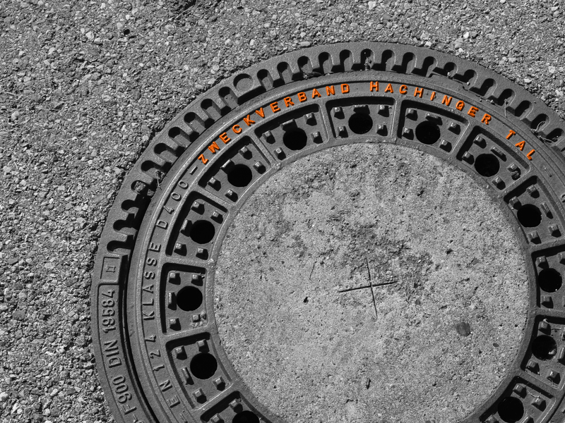 Ein runder Kanaldeckel mit der Aufschrift "Zweckverband Hachinger Tal" liegt auf einem körnigen Asphaltboden.