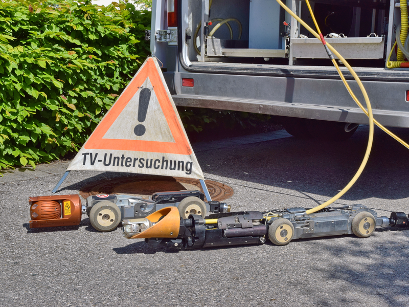Ein Warnschild mit der Aufschrift "TV-Untersuchung" steht neben einer mobilen Kanal-Inspektionseinheit und Kabeln, die aus einem Fahrzeug herausragen.