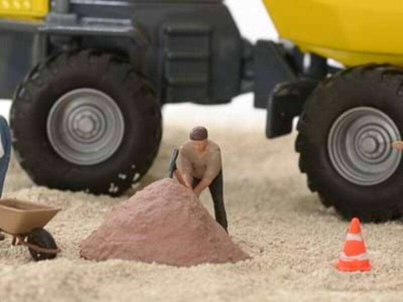 Miniaturfiguren von Bauarbeitern mit Werkzeugen und Verkehrsleitkegeln stehen vor einem großen Spielzeug-Lastwagen auf sandigem Untergrund.